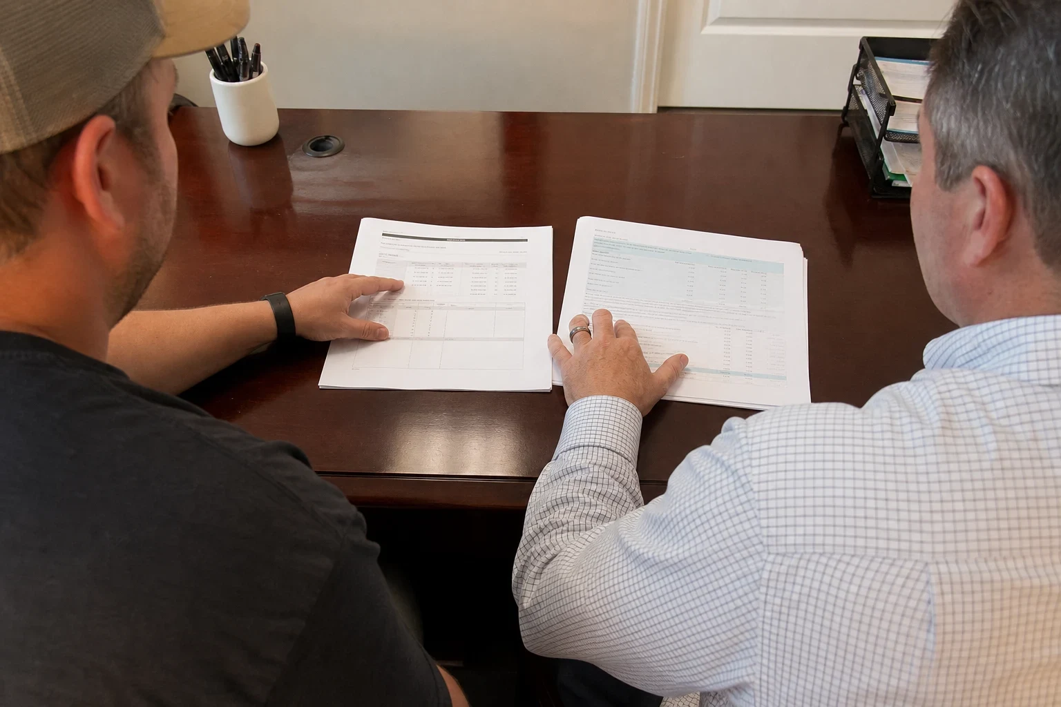 Over-the-shoulder view of two men at a wooden office desk reviewing two printed documents side by side, with a pen cup and paper tray nearby.