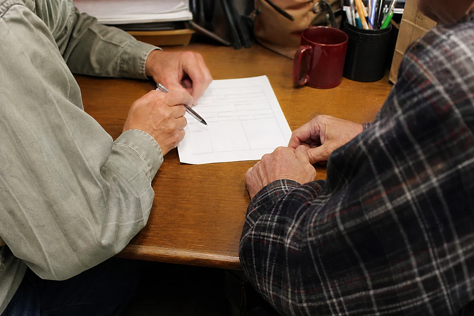 Two people at a wooden desk, one pointing a pen at a short form while an older man in a plaid flannel shirt rests his hands nearby; mug and pen cup in back.